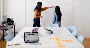 Two women stand in front of a whiteboard covered in writing and orange Post-Its,.