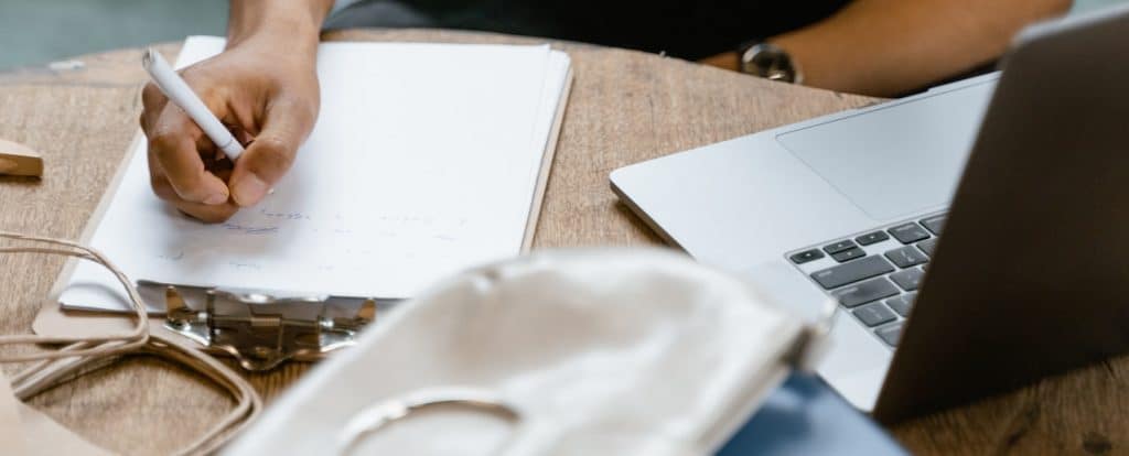 A man holds a pen over a clipboard, which is sitting beside a laptop on a round table.