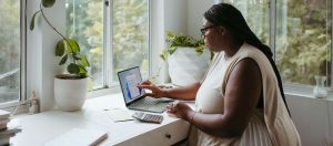 A woman sits at a desk, windows beyond showing lots of greenery. A laptop is open in front of her.