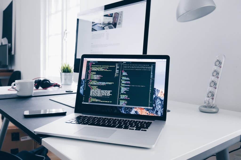 a laptop on a desk, a monitor behind. The laptop shows a code writing program in progress