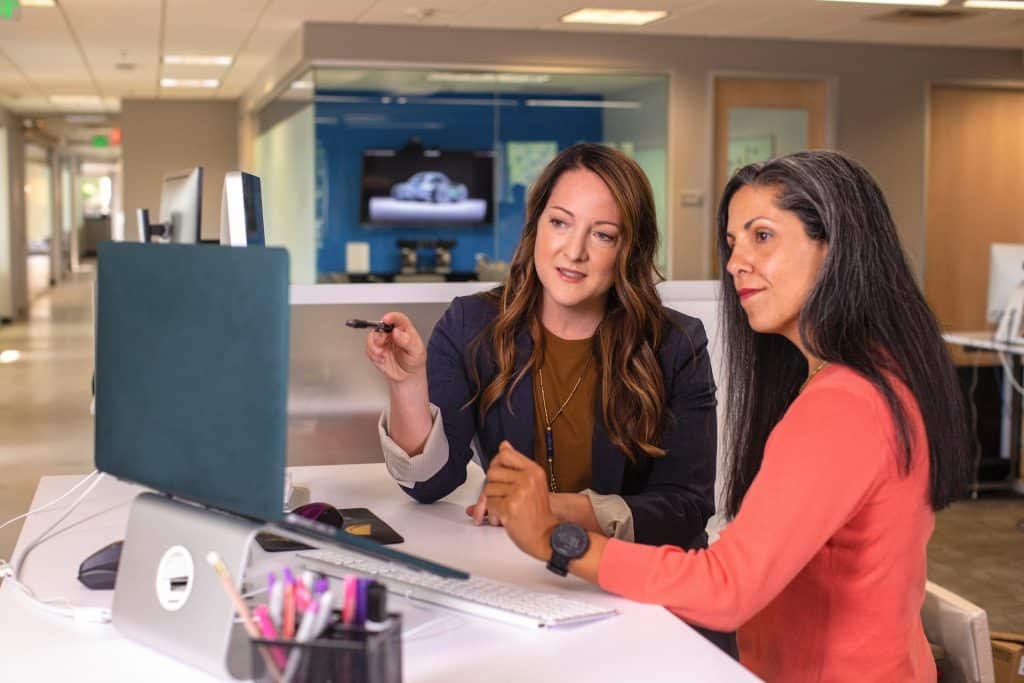 Two women sit on the same side of an office desk, both looking at a computer monitor.