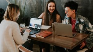 Three coworkers sit around a tall table, laptops open, laughing.