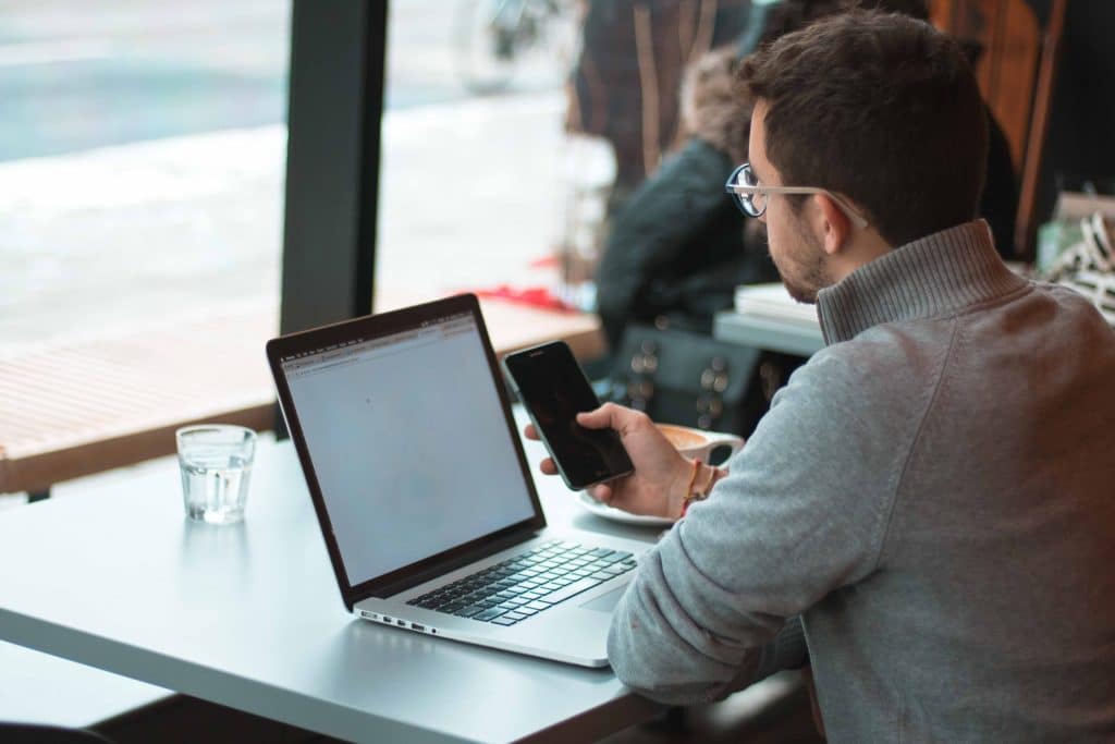 A man sits at a cafe table, his back to us. He is holding an iphone next to his open laptop.