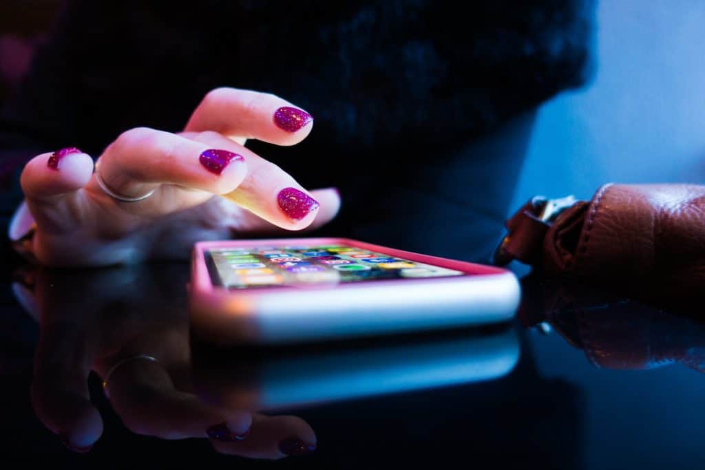 A hand with sparkly pink nail polish hovers above apps on a smartphone. The lighting is moody.