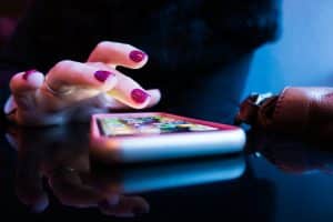 A hand with sparkly pink nail polish hovers above apps on a smartphone. The lighting is moody.