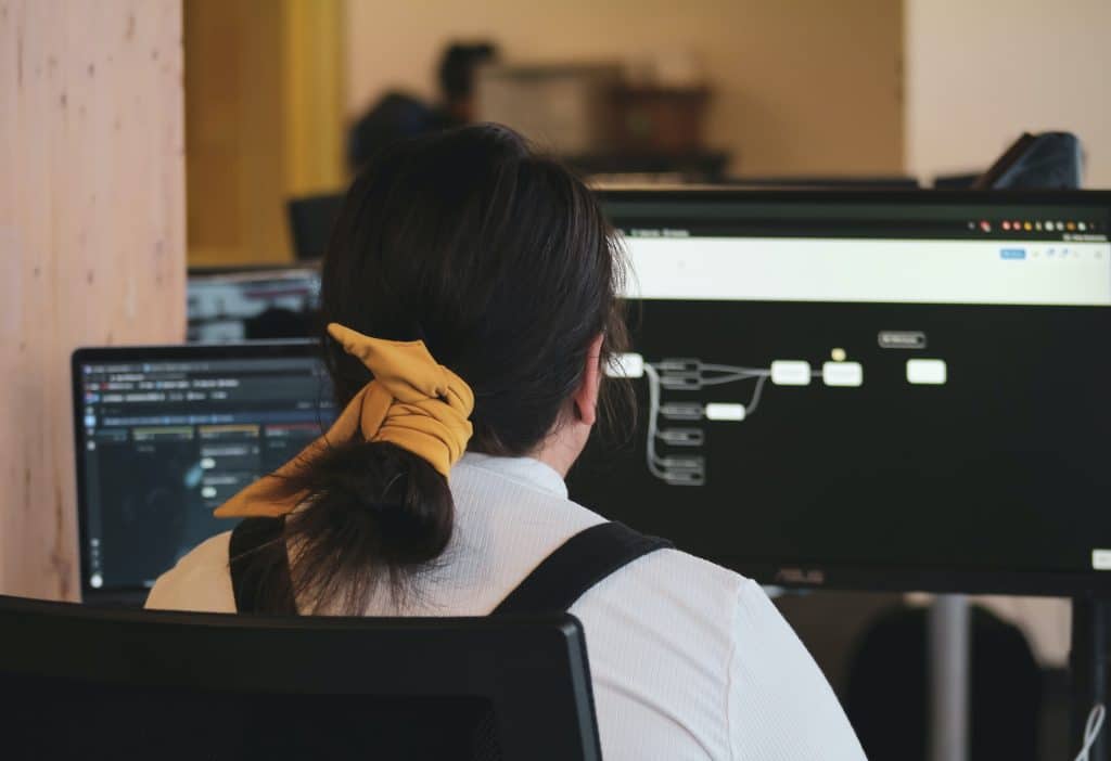 A woman with her hair ties in a yellow scarf sits with her back to the camera, a dual computer screen in front of her. The left screen shows code, the right shows a workflow.