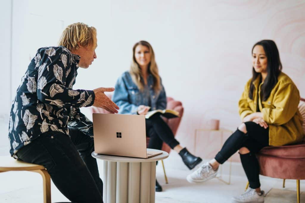 A man presents from a laptop, facing two women.