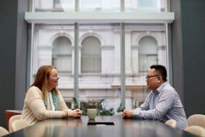 Anna and Hai sit opposite each other across a table, a large window behind them.