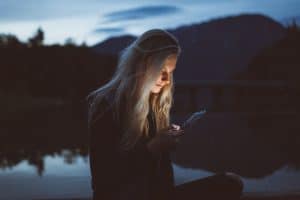 A woman stands in half darkness, a lake behind her. Her face is illuminated by a smartphone using Power Apps offline.