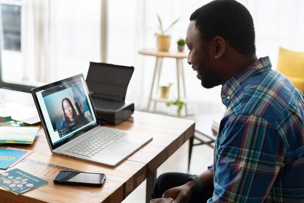 A man sits at a desk, looking at his laptop and smiling. A woman's face is on the screen, they are video calling.