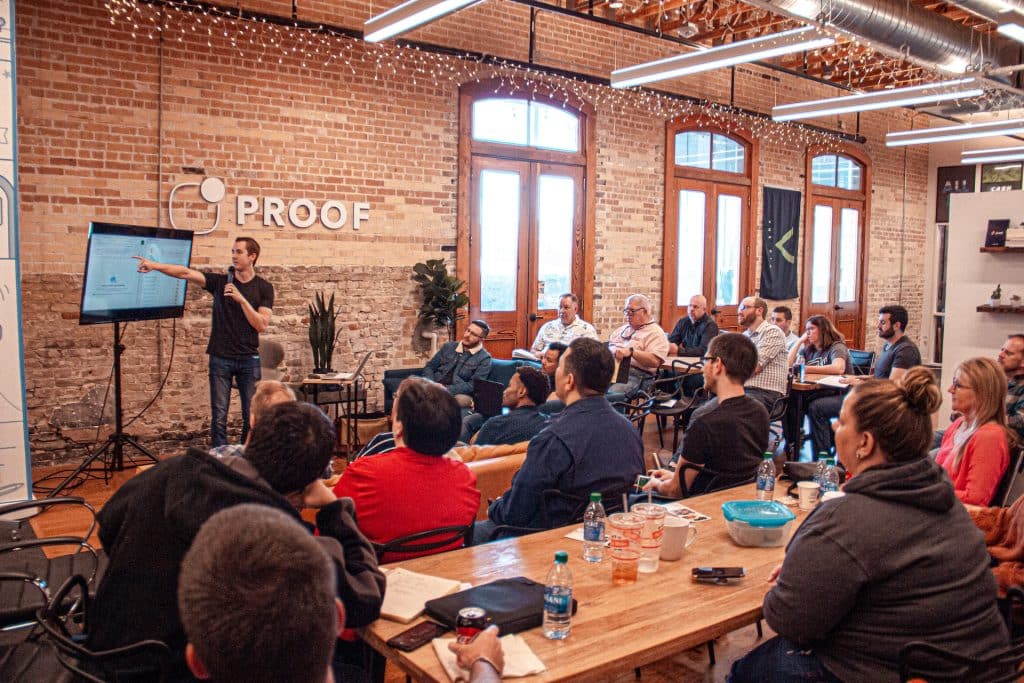 A large brick office, long timber tables. A team meeting in progress, one worker holding a mic and pointing to a screen