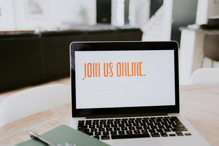 An open laptop on a desk, a bright window beyond. The screen is white with orange writing: JOIN US ONLINE