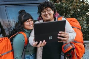 A man and a woman stand in front of a van, high-vis orange travel packs on their back. They smile at a Microsoft tablet held aloft in front of them.