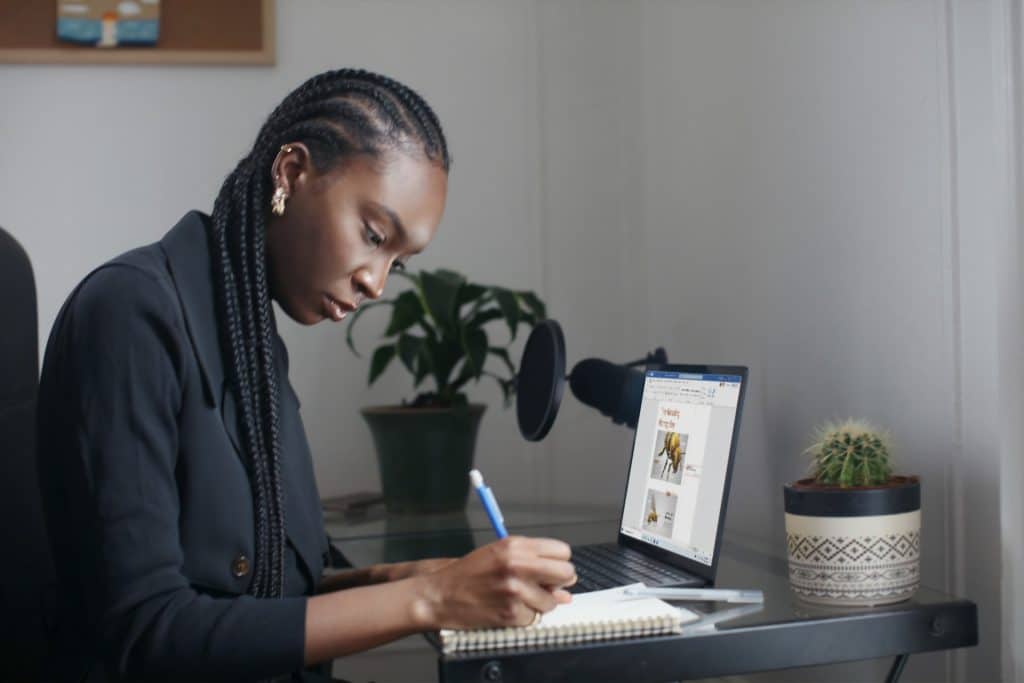 A woman sits at her home office desk, her laptop open. She is making a note on a notepad.