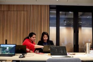 Two women sit side by side, one points at a laptop to define the channels in Microsoft Teams