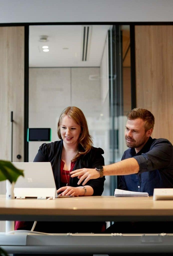 A male and female colleague sit behind a laptop, the man gesturing towards the screen at document management