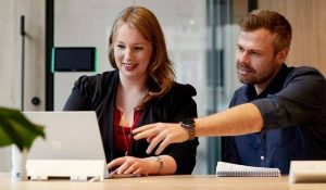 A male and female colleague sit behind a laptop, the man gesturing towards the screen at document management