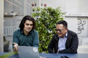 Two men sit at an outside bench gesturing towards a laptop in front of them, discussing intranet content management