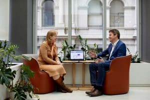 Two colleague sit facing each other in front of a large window, a laptop on the table between them, discussing change