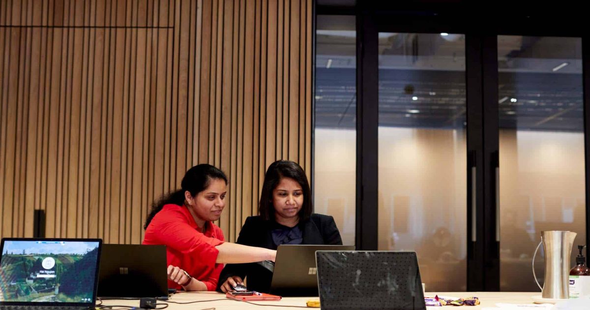 Two women sit side by side, one points at a laptop to define the channels in Microsoft Teams