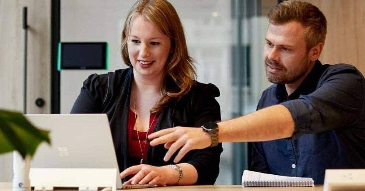A male and female colleague sit behind a laptop, the man gesturing towards the screen at document management