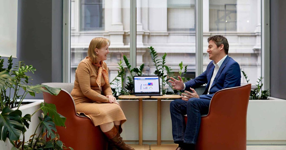 Two colleague sit facing each other in front of a large window, a laptop on the table between them, discussing change