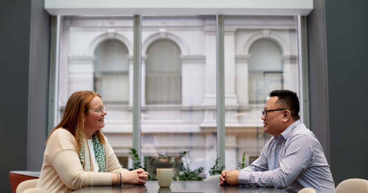 Anna and Hai sit opposite each other across a table, a large window behind them.