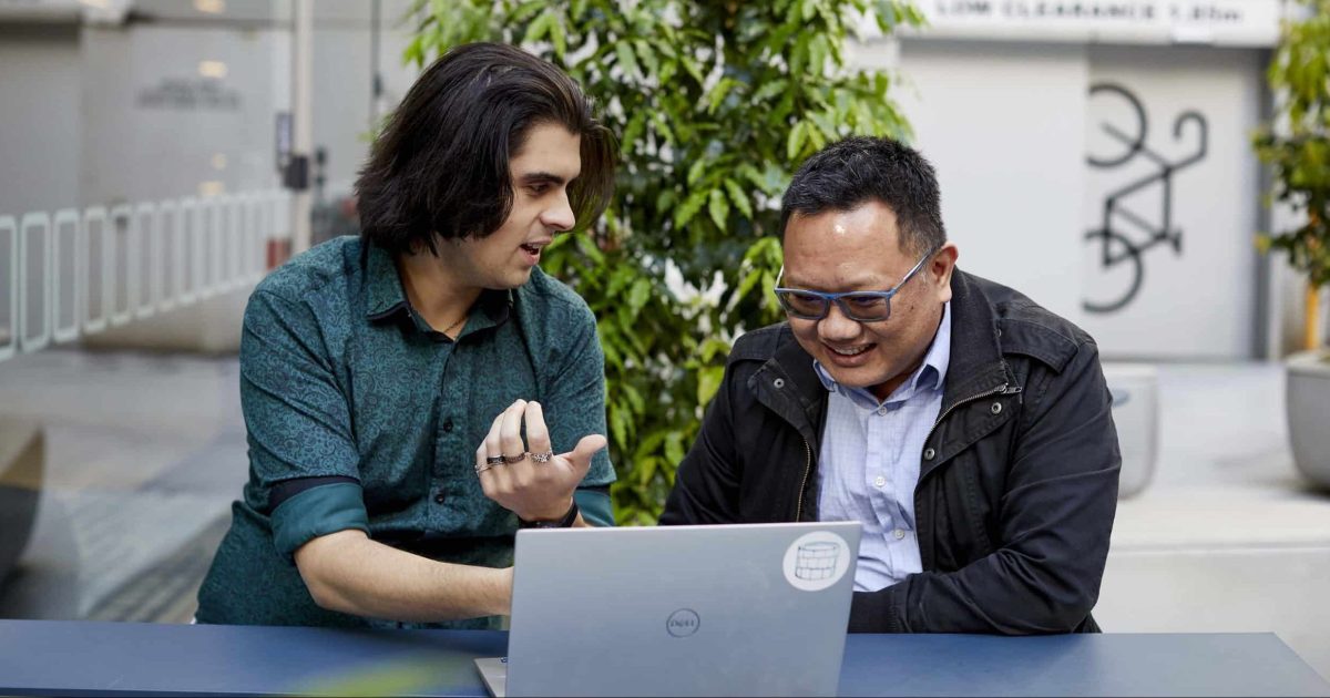 Two men sit side by side discussing Power Bi visualisation tricks at an outdoor table with a laptop