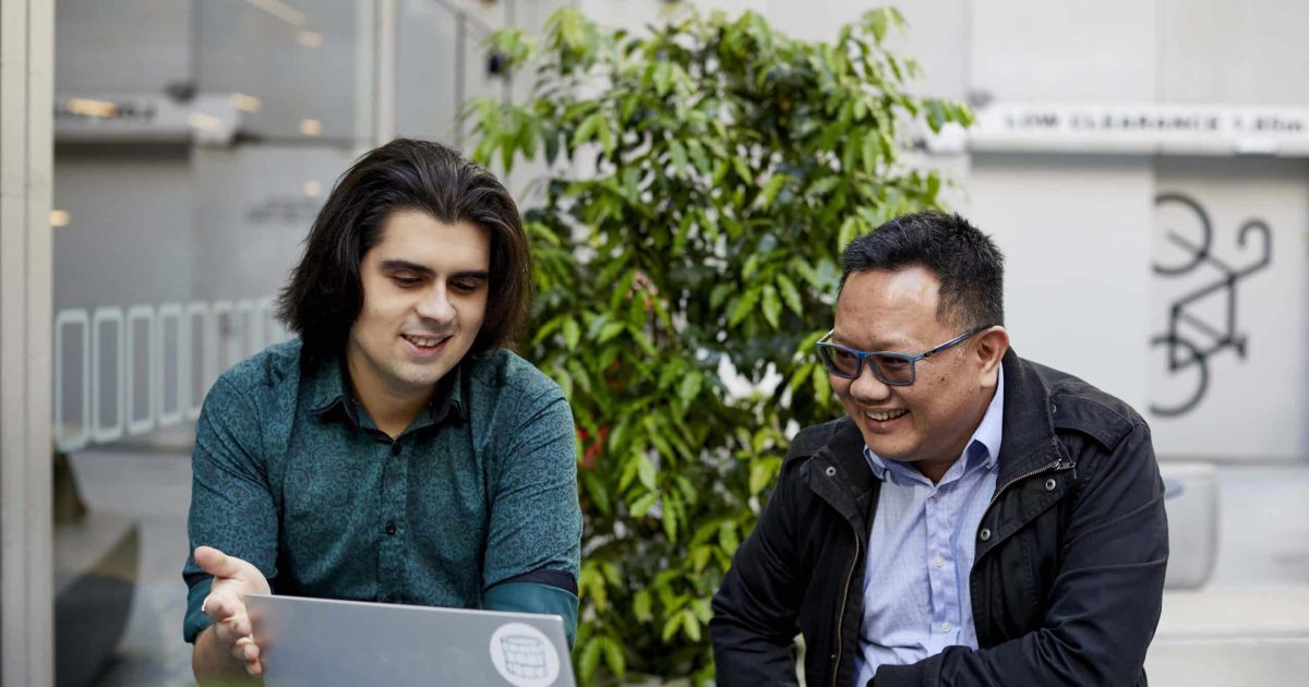 Two men sit at an outside bench gesturing towards a laptop in front of them, discussing intranet content management