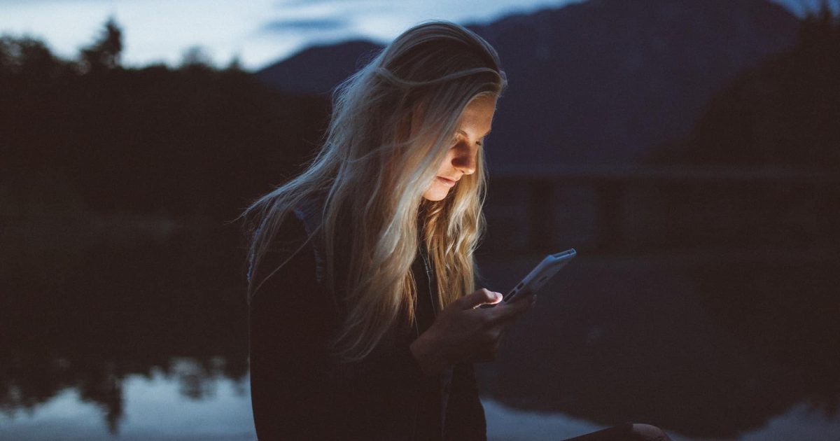 A woman stands in half darkness, a lake behind her. Her face is illuminated by a smartphone using Power Apps offline.