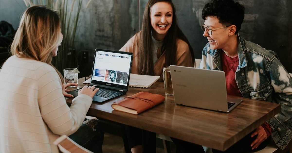 Three coworkers sit around a tall table, laptops open, laughing.