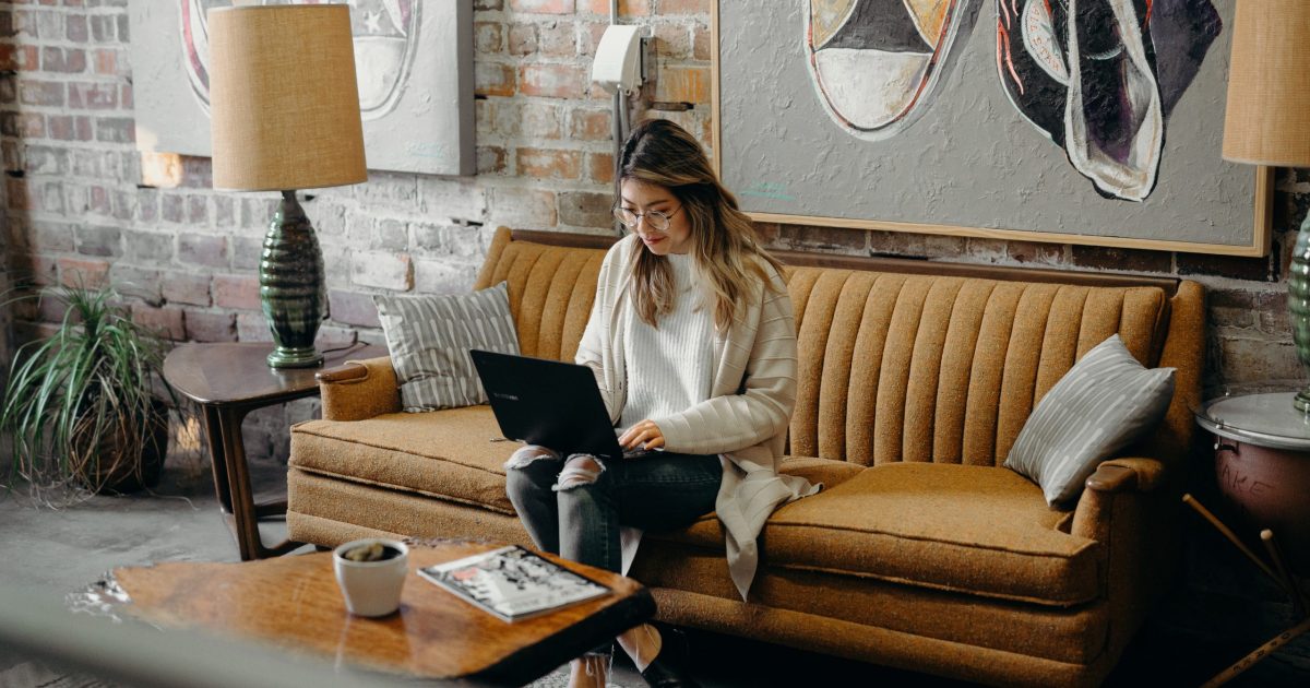 A woman sits on a yellow couch in a bright, quirky sitting area. She smiles at the laptop on her lap.