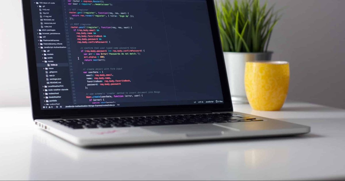 A silver laptop is open on a desk, a mug and small grassy plant behind to the right. On the screen is a coding script.