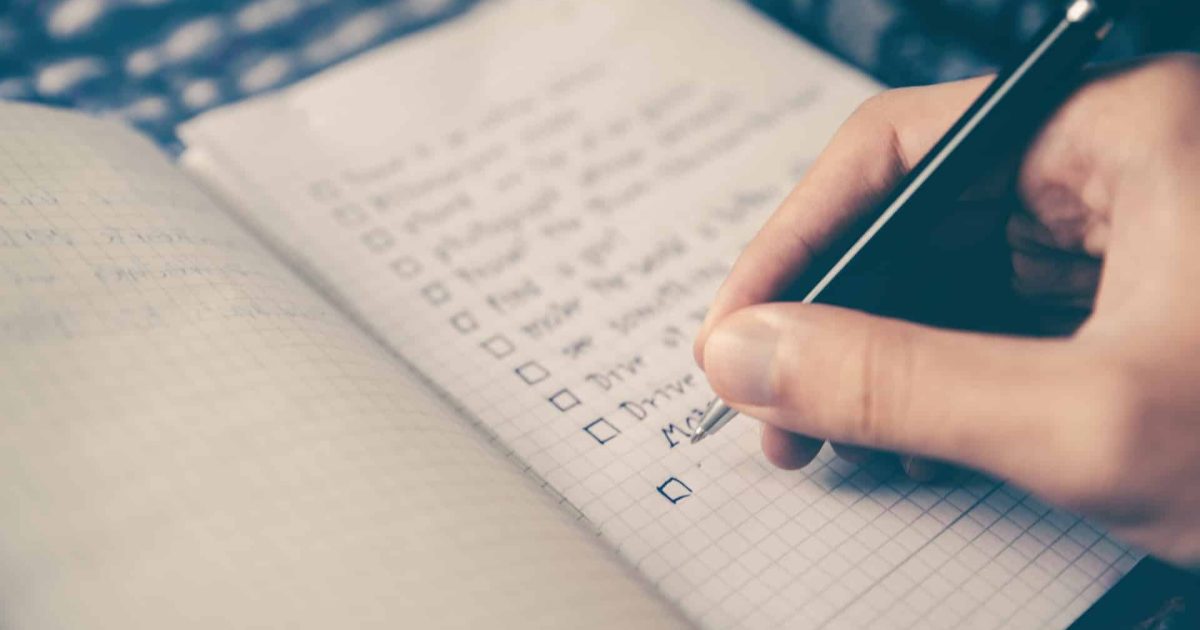 An open notebook, on the right hand side is a long check list with boxes in the left-hand margin. A hand with a pen is in the bottom right corner.