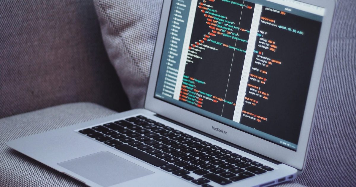 A laptop sits on the arm of a grey couch, a phone beside it. The computer shows a code running.