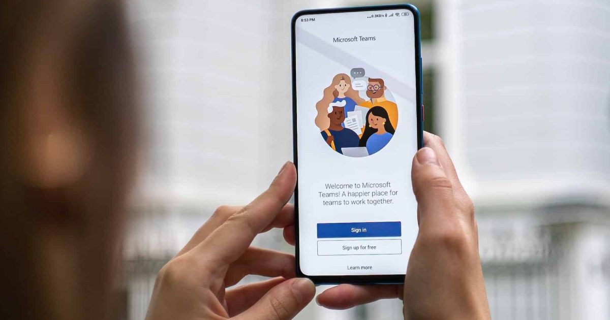 A pair of male hands hold a smartphone aloft, a window in the background. On the screen is the log in screen of Microsoft Teams.