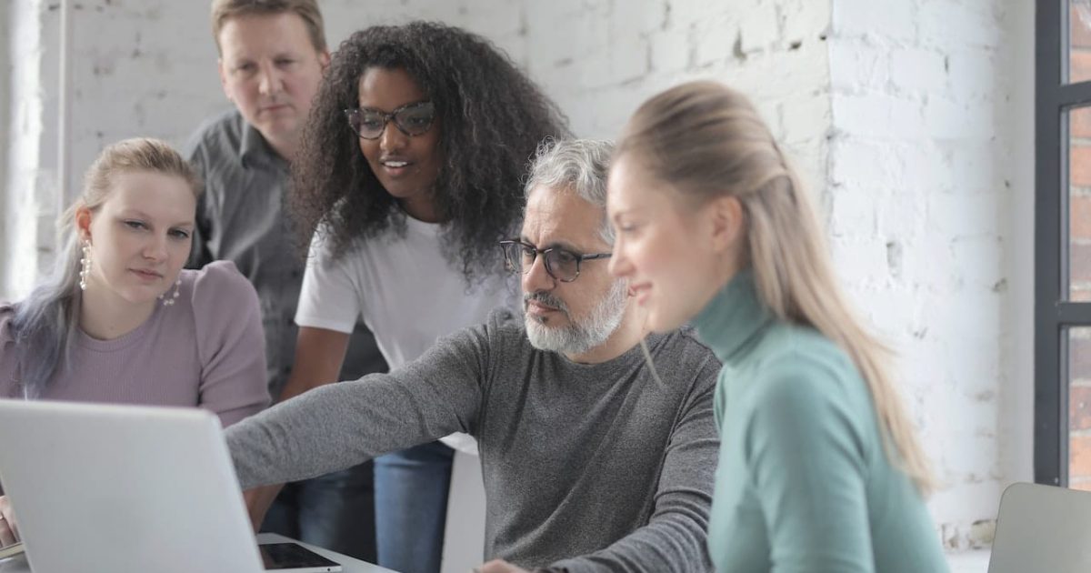 Five people gather around a laptop smiling as one of them points to the screen.