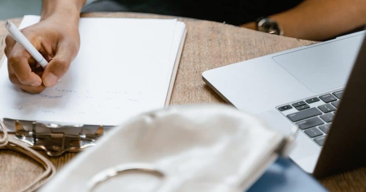 A man holds a pen over a clipboard, which is sitting beside a laptop on a round table.