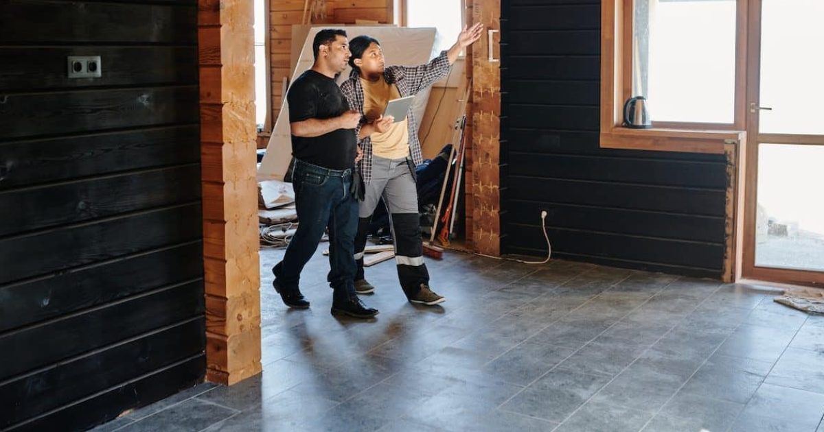 A man and a woman walk through a room, wearing building attire. They are comparing something to a design.