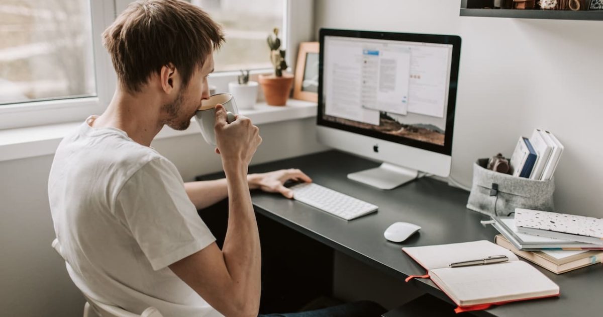 A man in a white shirt sips from a mug, sitting at a desk. A computer screen and open notebooks are on the desk.