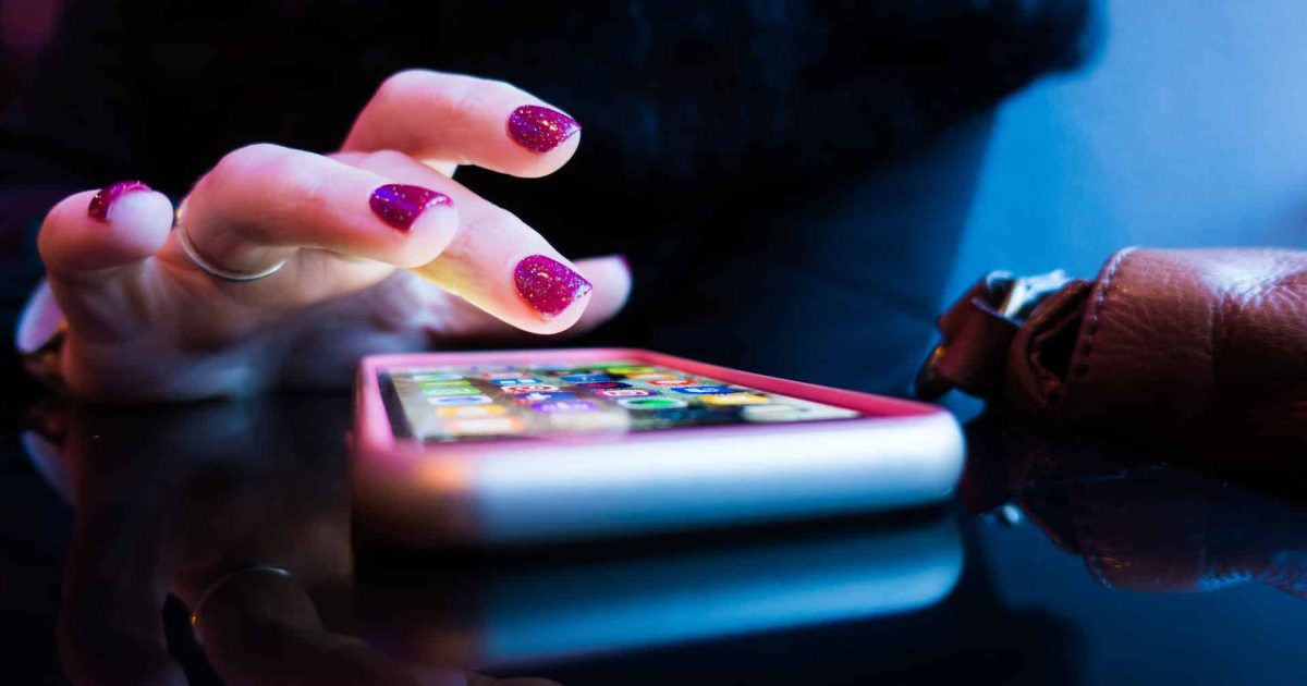 A hand with sparkly pink nail polish hovers above apps on a smartphone. The lighting is moody.