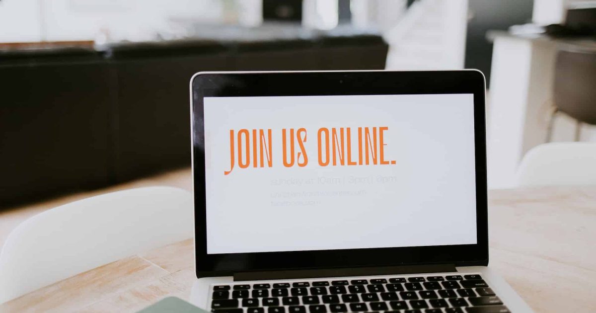An open laptop on a desk, a bright window beyond. The screen is white with orange writing: JOIN US ONLINE