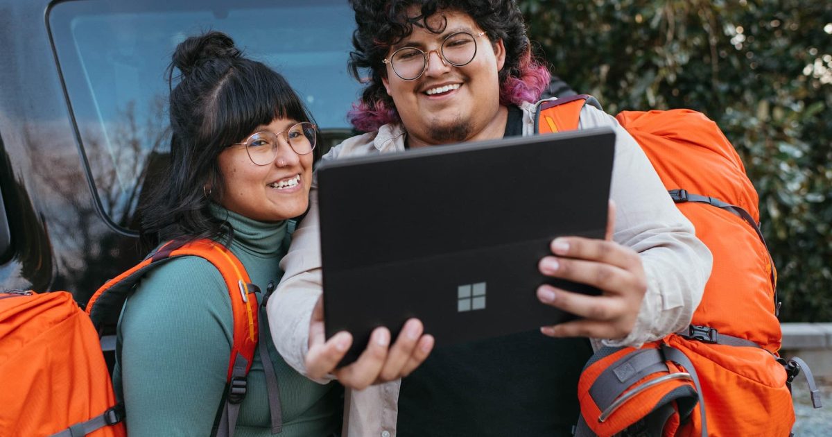 A man and a woman stand in front of a van, high-vis orange travel packs on their back. They smile at a Microsoft tablet held aloft in front of them.