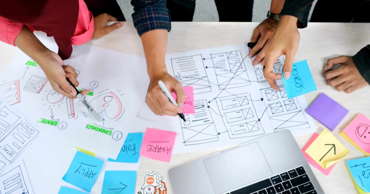 An overhead shot of a desk covered in colourful notes, and a hand-drawn map of a webpage. Different pairs of hands hold pens over the papers.