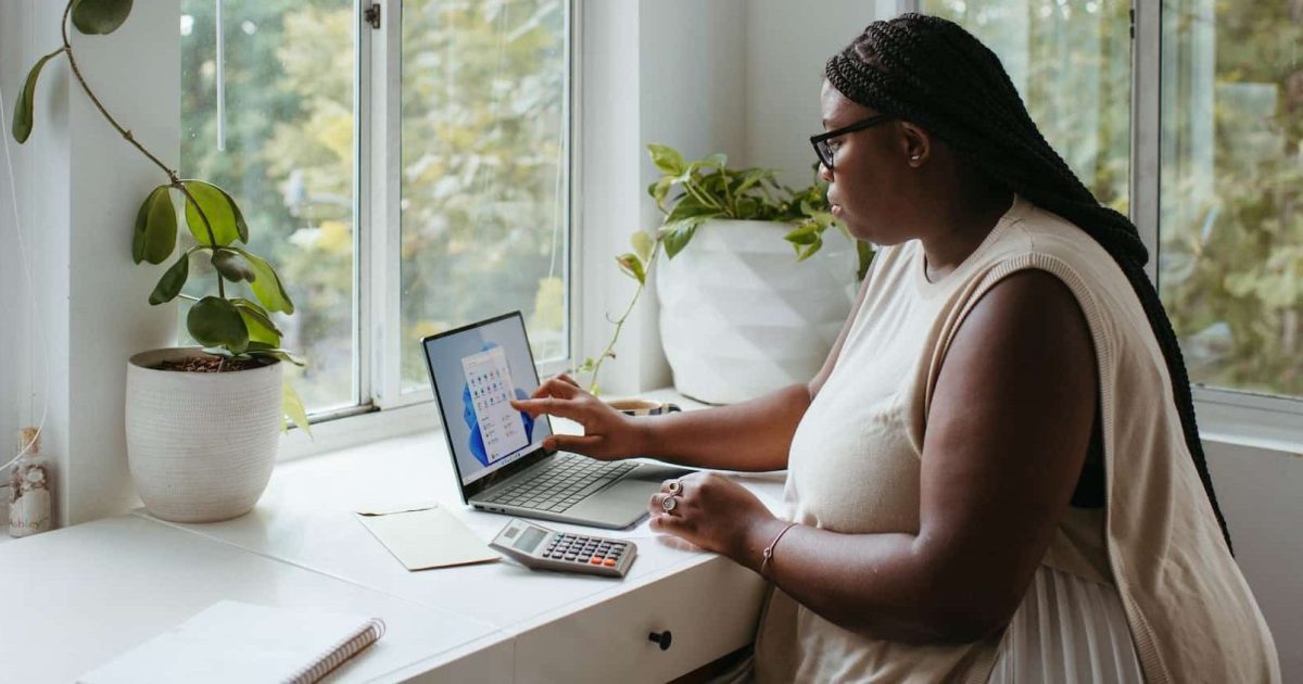 A woman sits at a desk, windows beyond showing lots of greenery. A laptop is open in front of her.